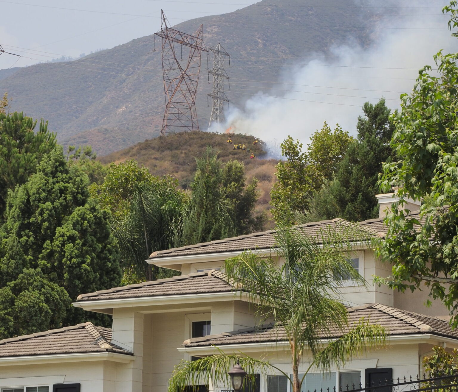 Photo of large home with wildfire and fire fighters in the hillside behind the home.